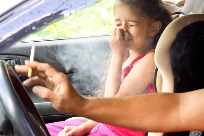 Child in car seat looks distressed by adult smoking in front, message on secondhand smoke dangers