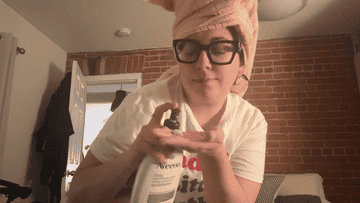 Person in glasses and towel on head holds a bottle, standing in a kitchen with brick wall