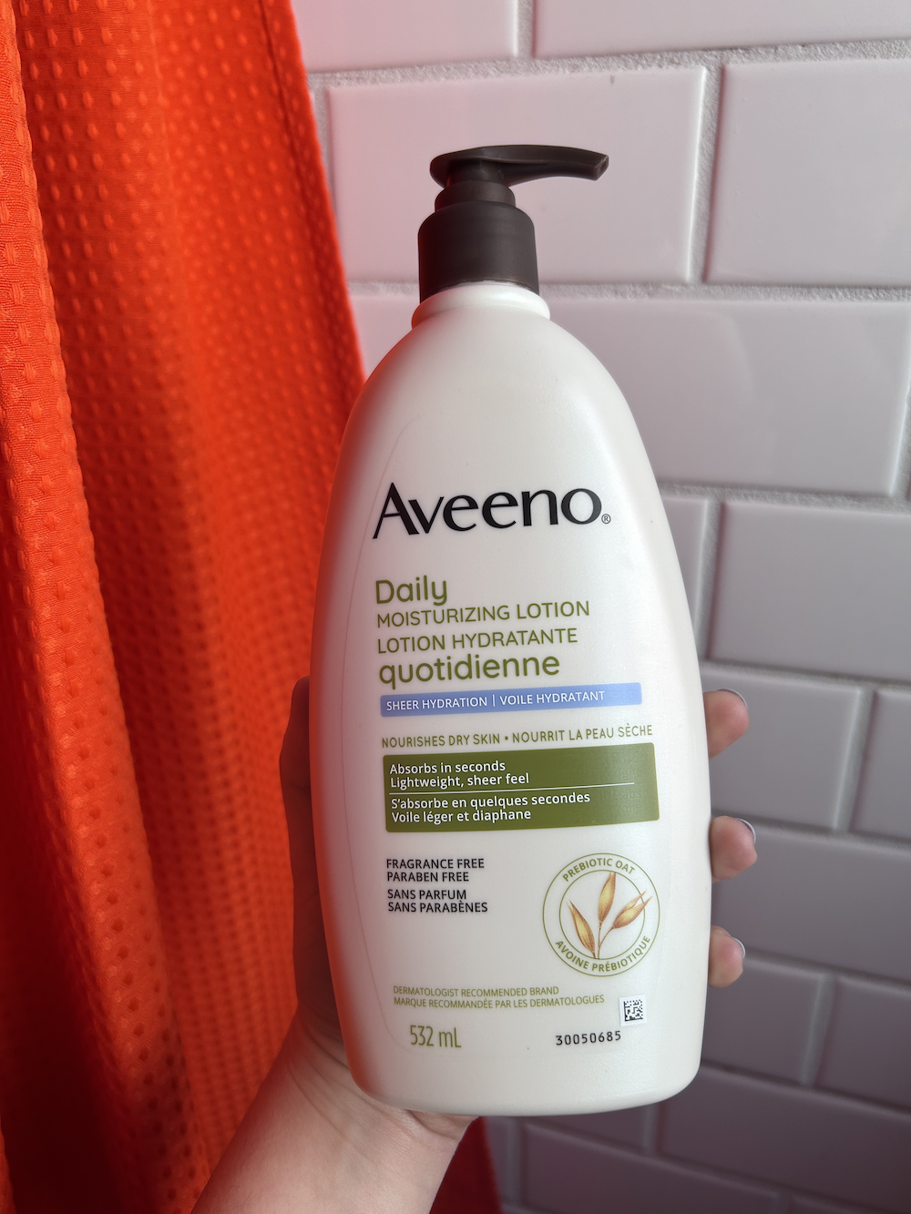 Hand holding a bottle of Aveeno Daily Moisturizing Lotion against an orange backdrop