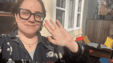 Person in glasses waving, with a sunny background and a wooden deck area