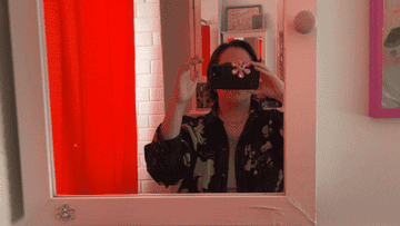 Person takes a selfie in mirror wearing a patterned jacket. Decor includes pink accents and reflective surfaces