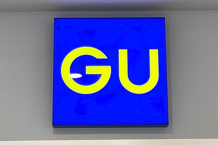 Sign with letters “GU” on blue background, mounted on a wall with ceiling lights visible