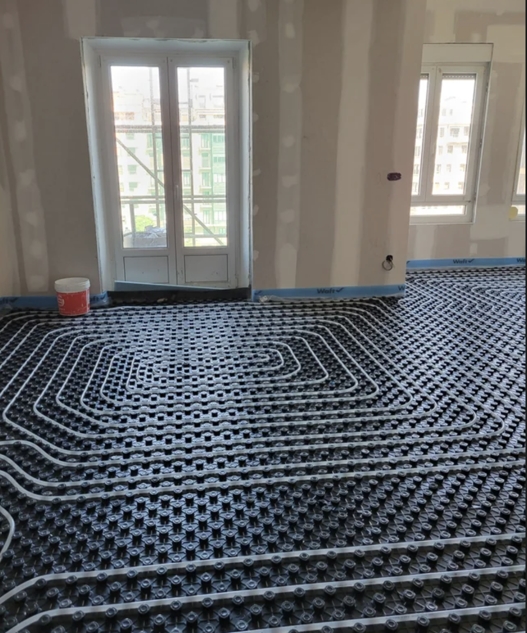 Intricate black floor tubing for underfloor heating installation in an unfurnished room with large windows and bare walls