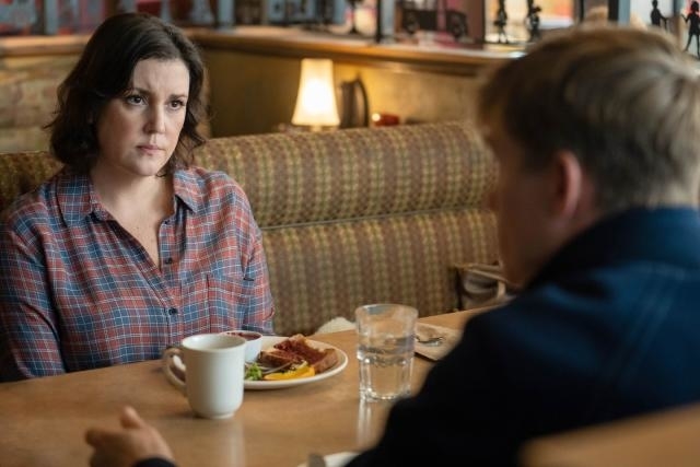 Actress in a plaid shirt looks intently at a person off-camera while seated at a diner table