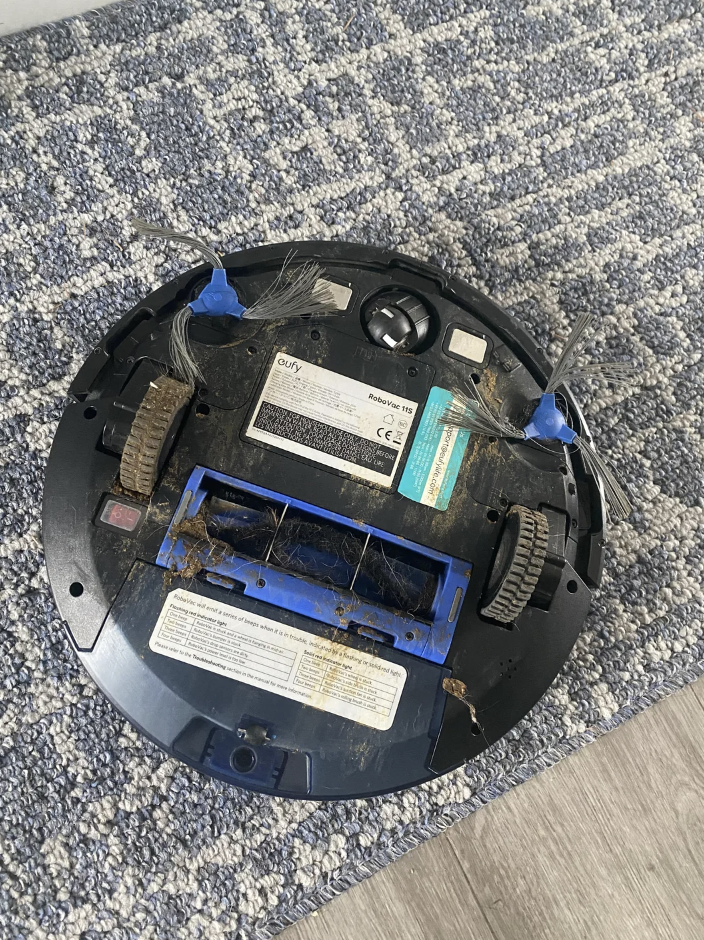 A damaged robot vacuum with debris and tangled hair on its brushes, lying on a carpeted floor