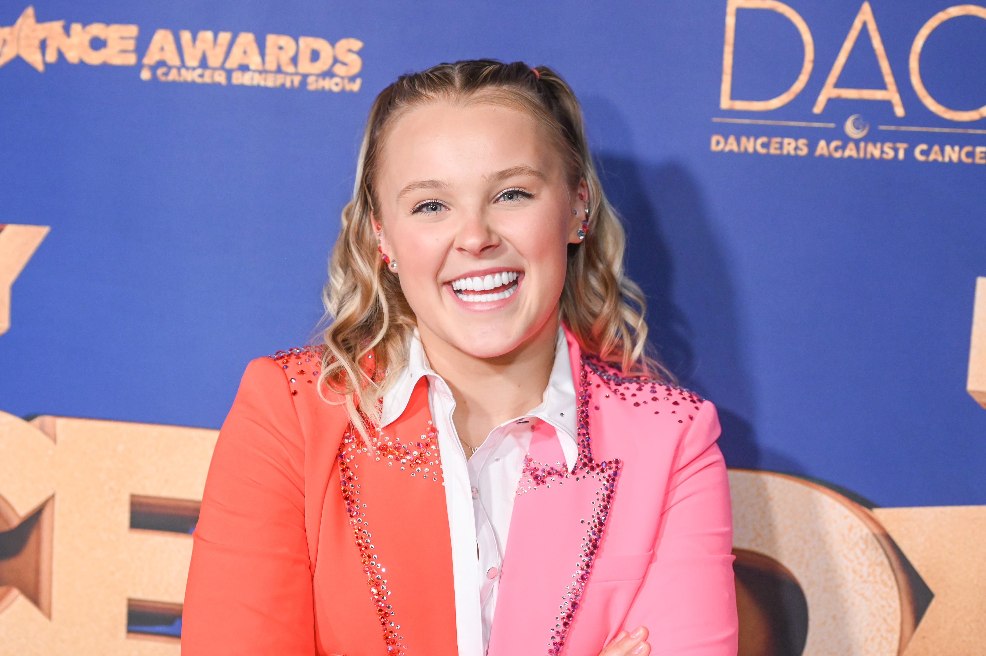JoJo Siwa smiling, wearing a pink shirt and orange jacket with details at a dance awards event
