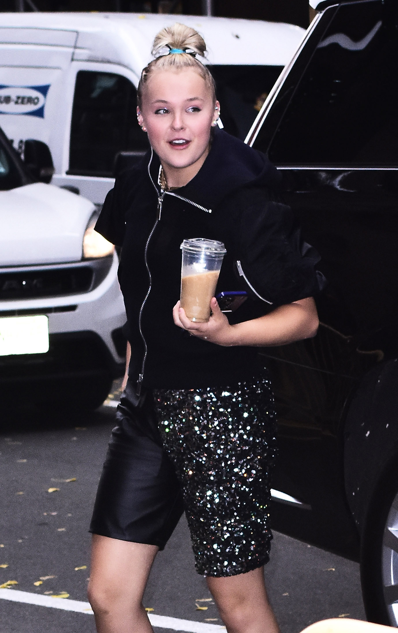 JoJo Siwa in a black jacket and sparkly skirt, holding a drink, walking on the street