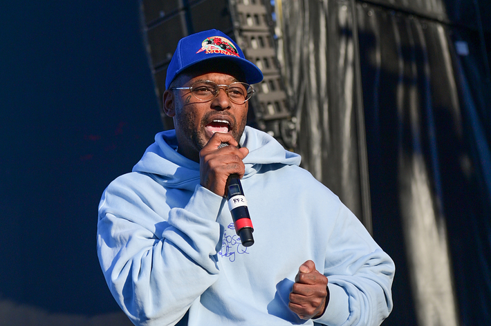 A person in a hoodie and baseball cap speaks into a microphone on stage
