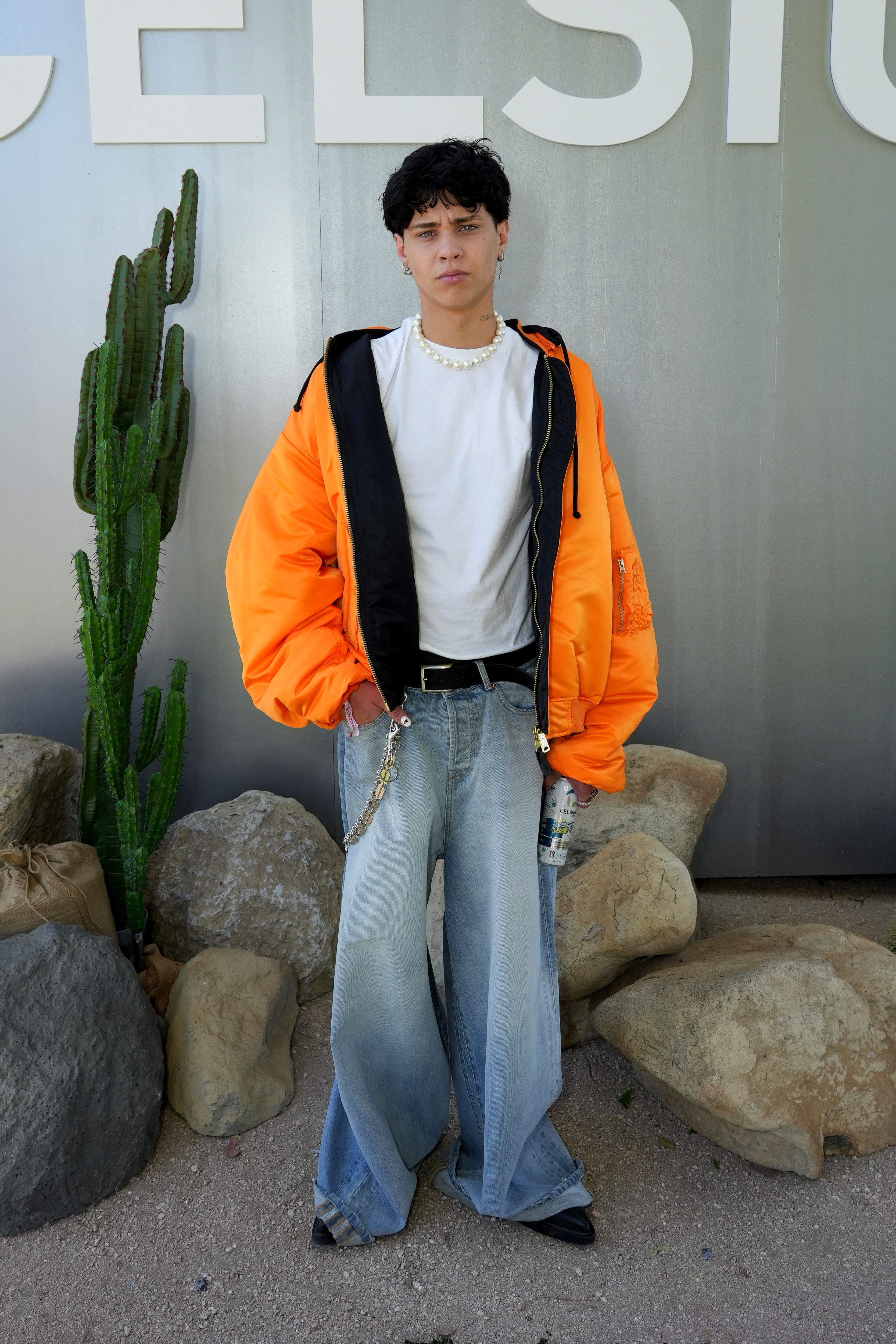 Person in an orange jacket, white top, wide-leg jeans, and a pearl necklace, standing in front of a cactus and rocks