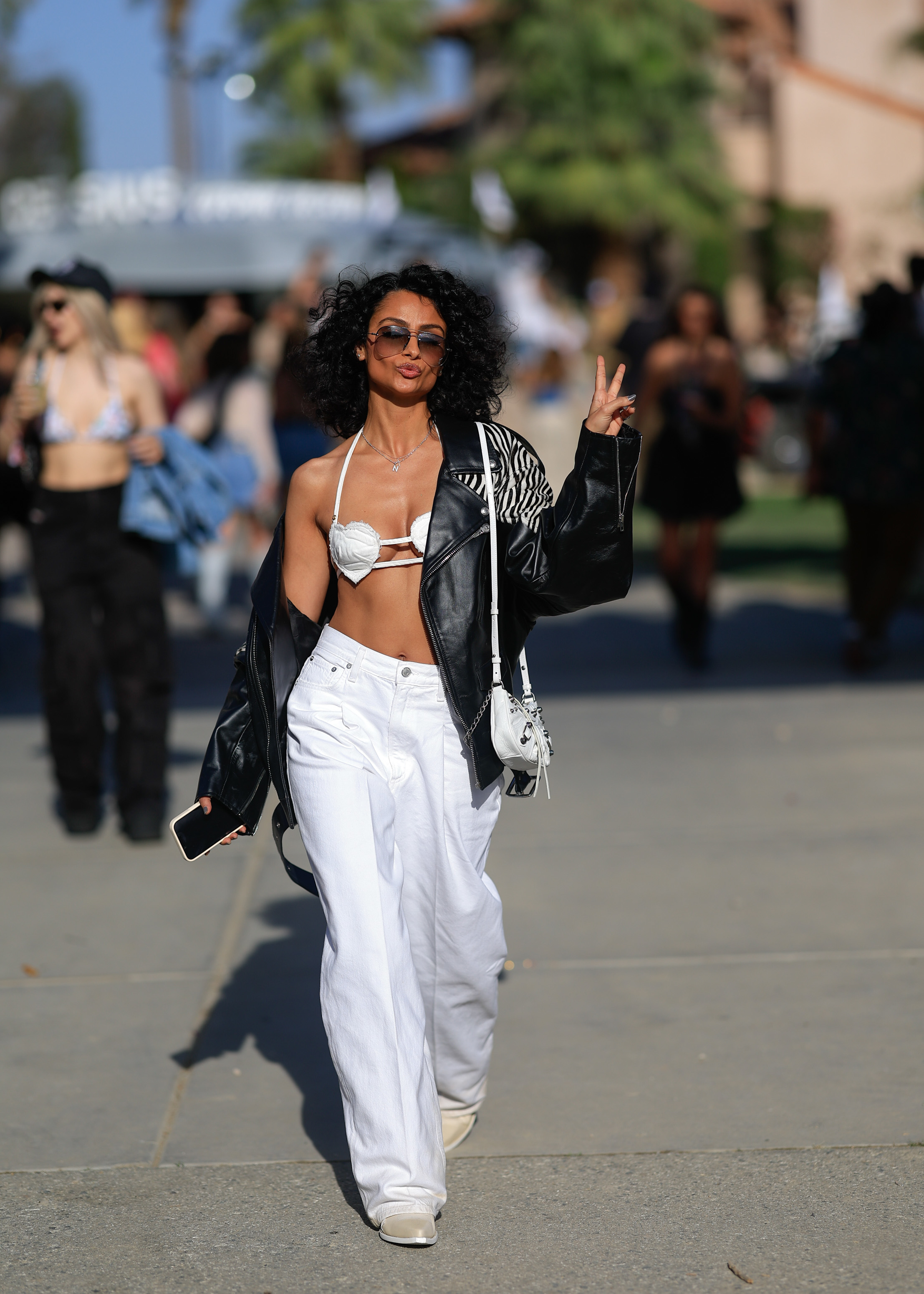 Woman in white pants, bralette, and black jacket makes peace sign, walking outdoors