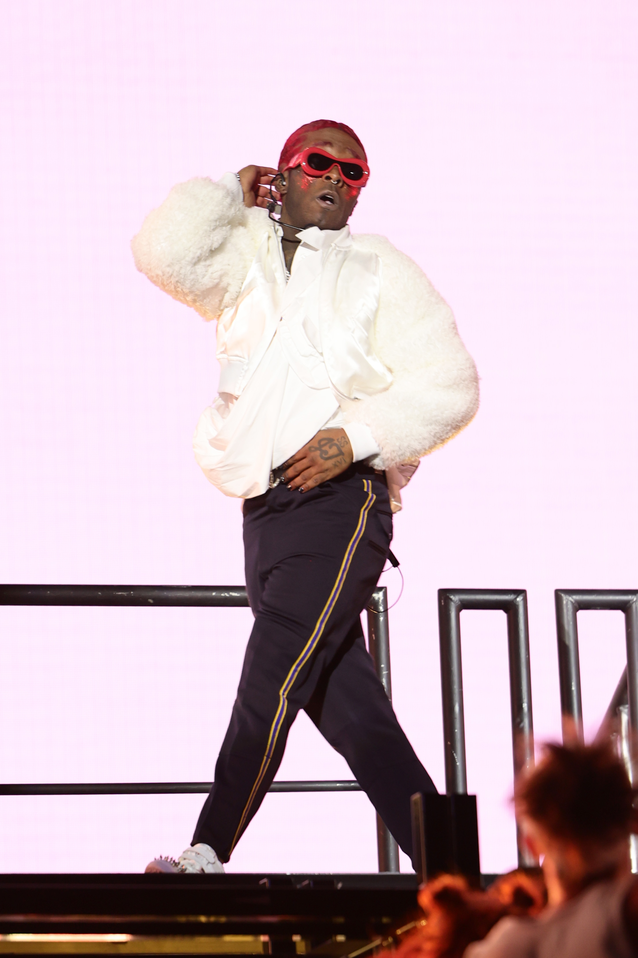 Performing artist on stage in a white fluffy jacket and striped pants, accessorized with red sunglasses