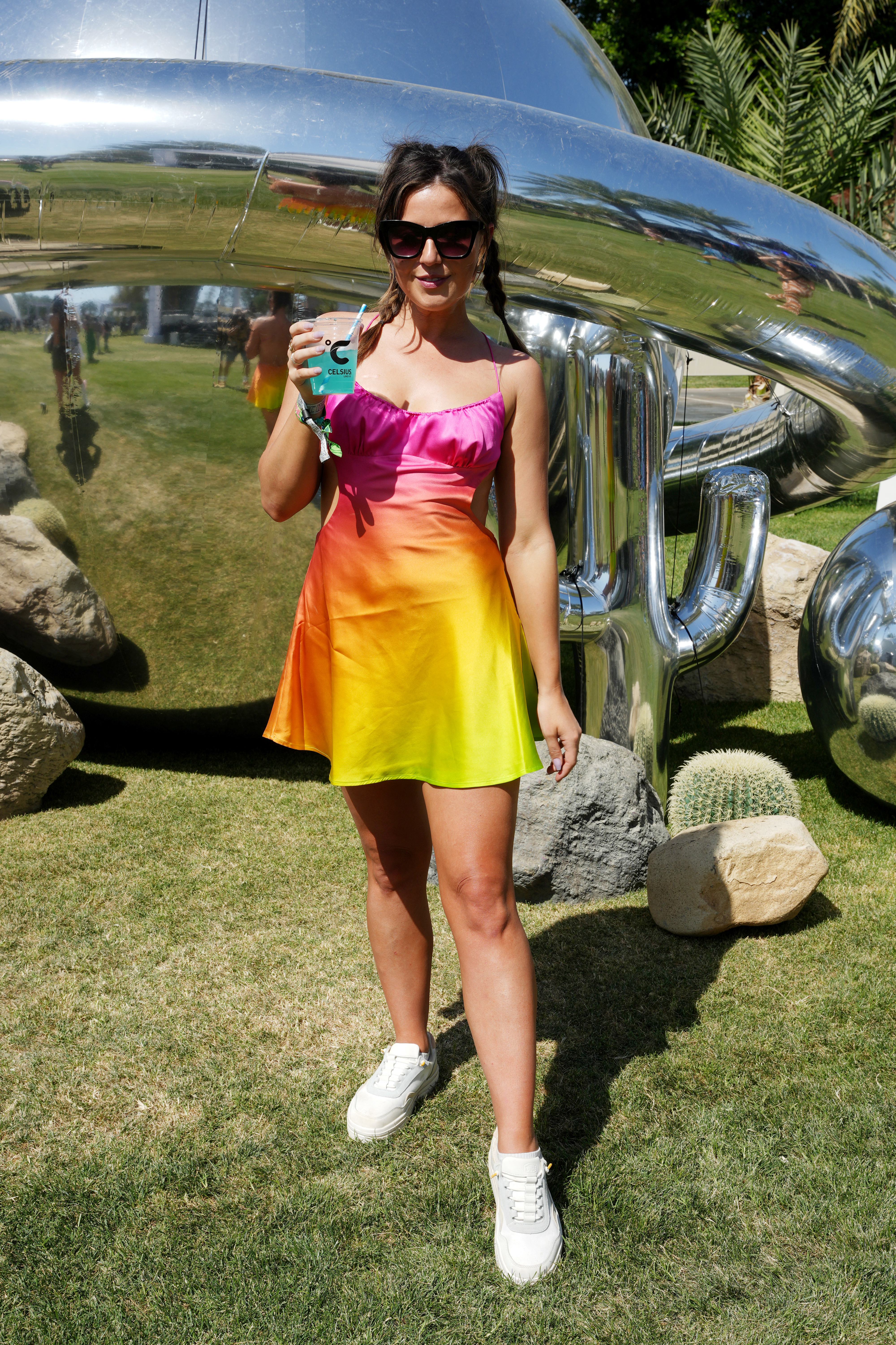 Woman in a colorful sleeveless dress and sneakers stands in front of a reflective sculpture, holding a beverage