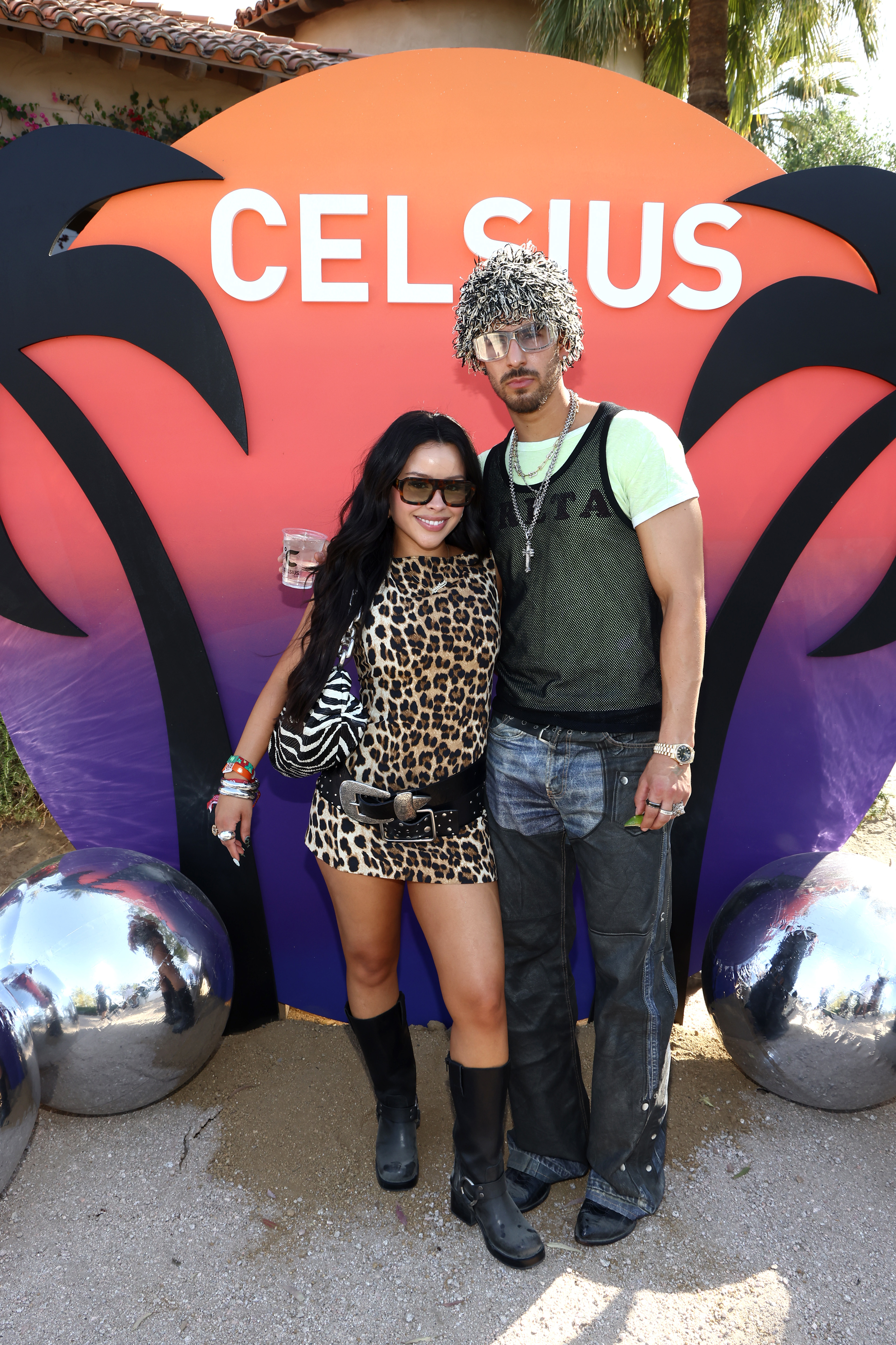 Two individuals standing before a Celsius logo, one in a leopard print outfit and boots, the other in a tank top and hat