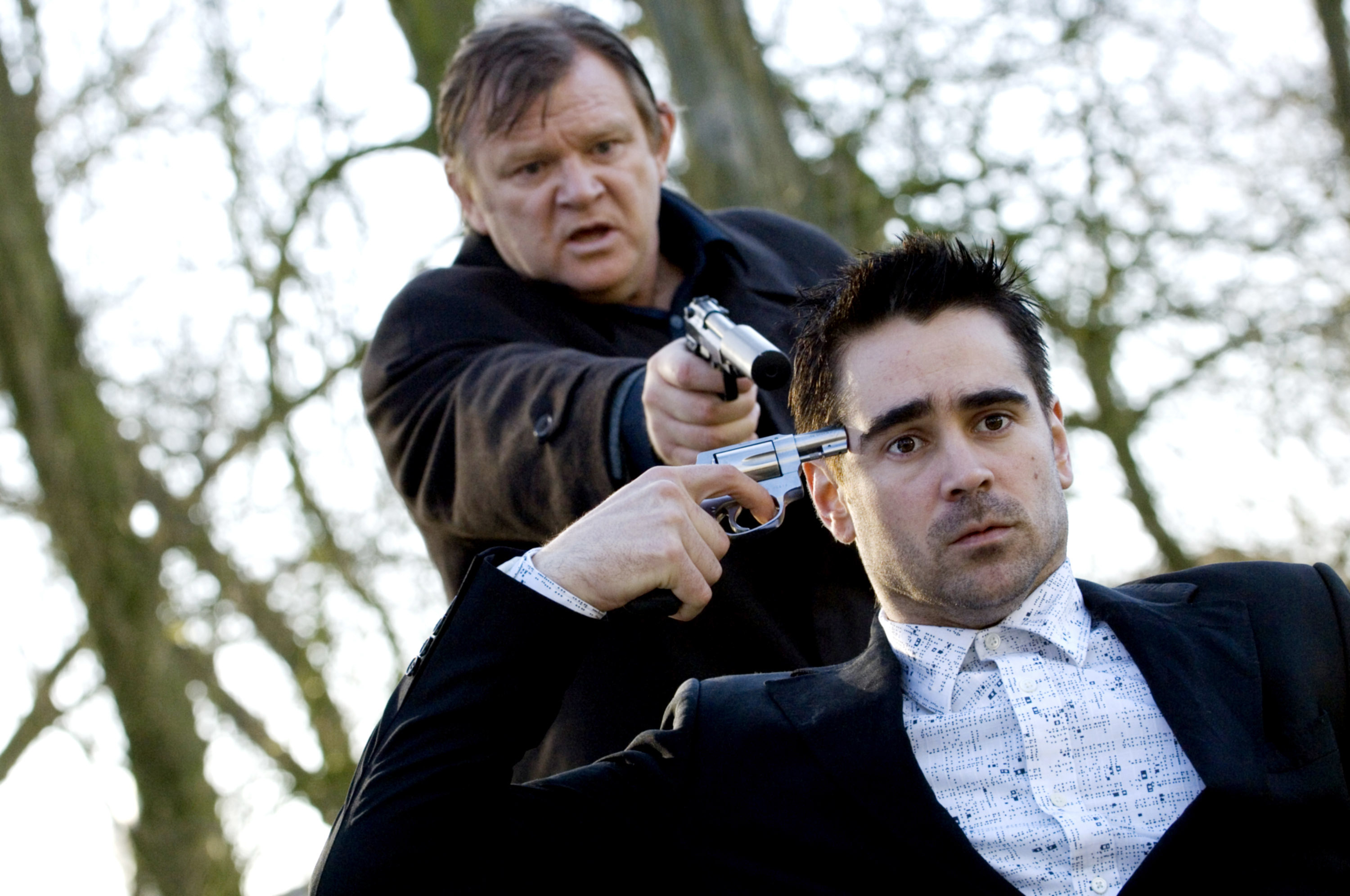 Two actors in a tense scene with one holding a gun to the other&#x27;s head, outdoors with trees in the background