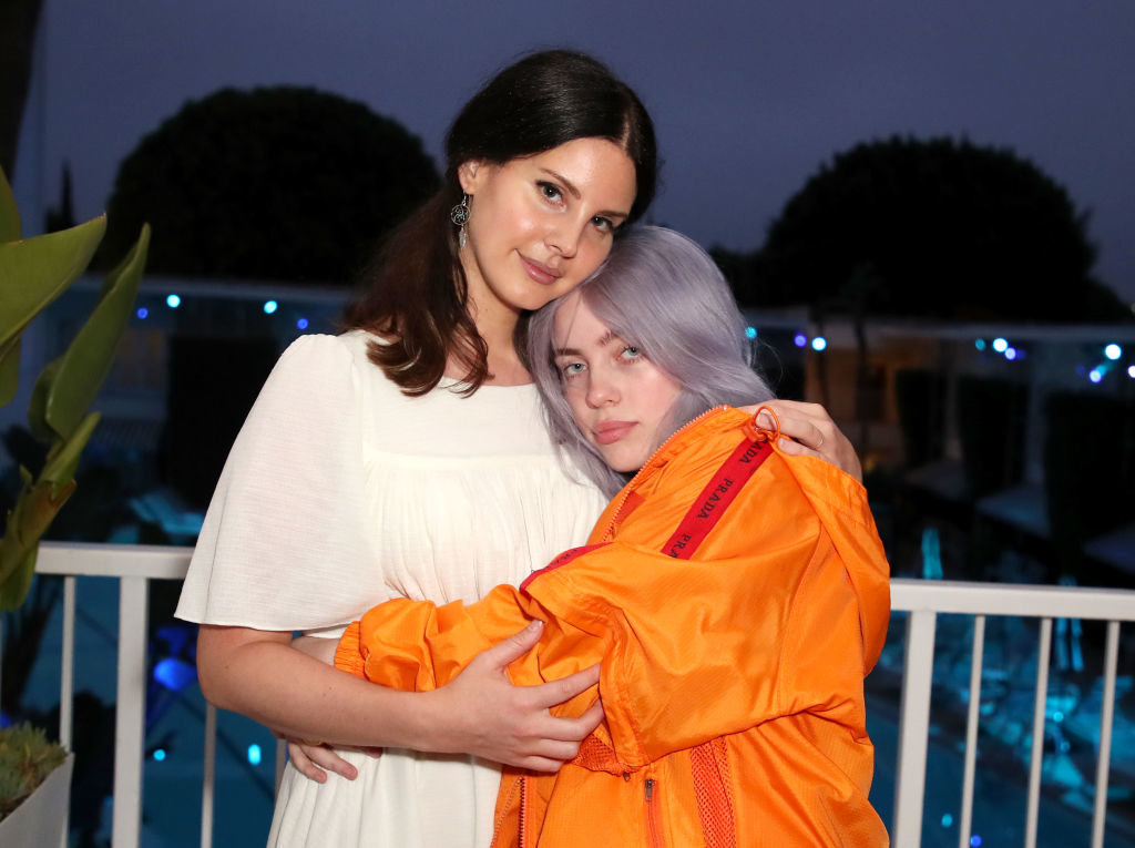 Lana Del Rey and Billie Eilish posing together, Del Rey in a white dress, Eilish in an orange jacket