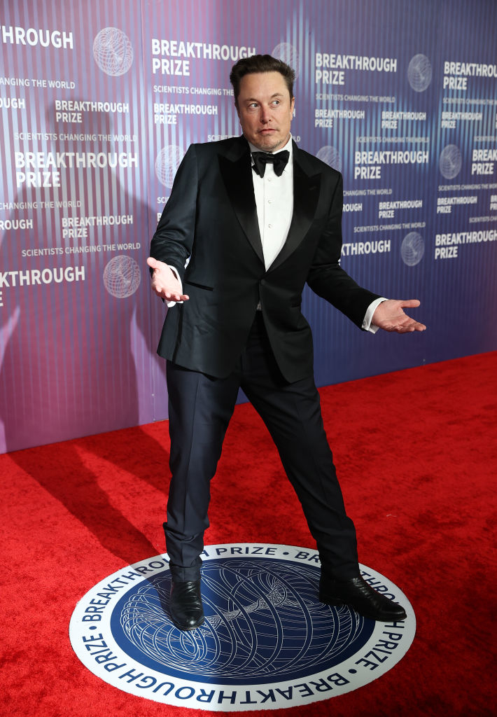 Elon Musk in a black tuxedo with bow tie, standing with hands outstretched at the Breakthrough Prize event
