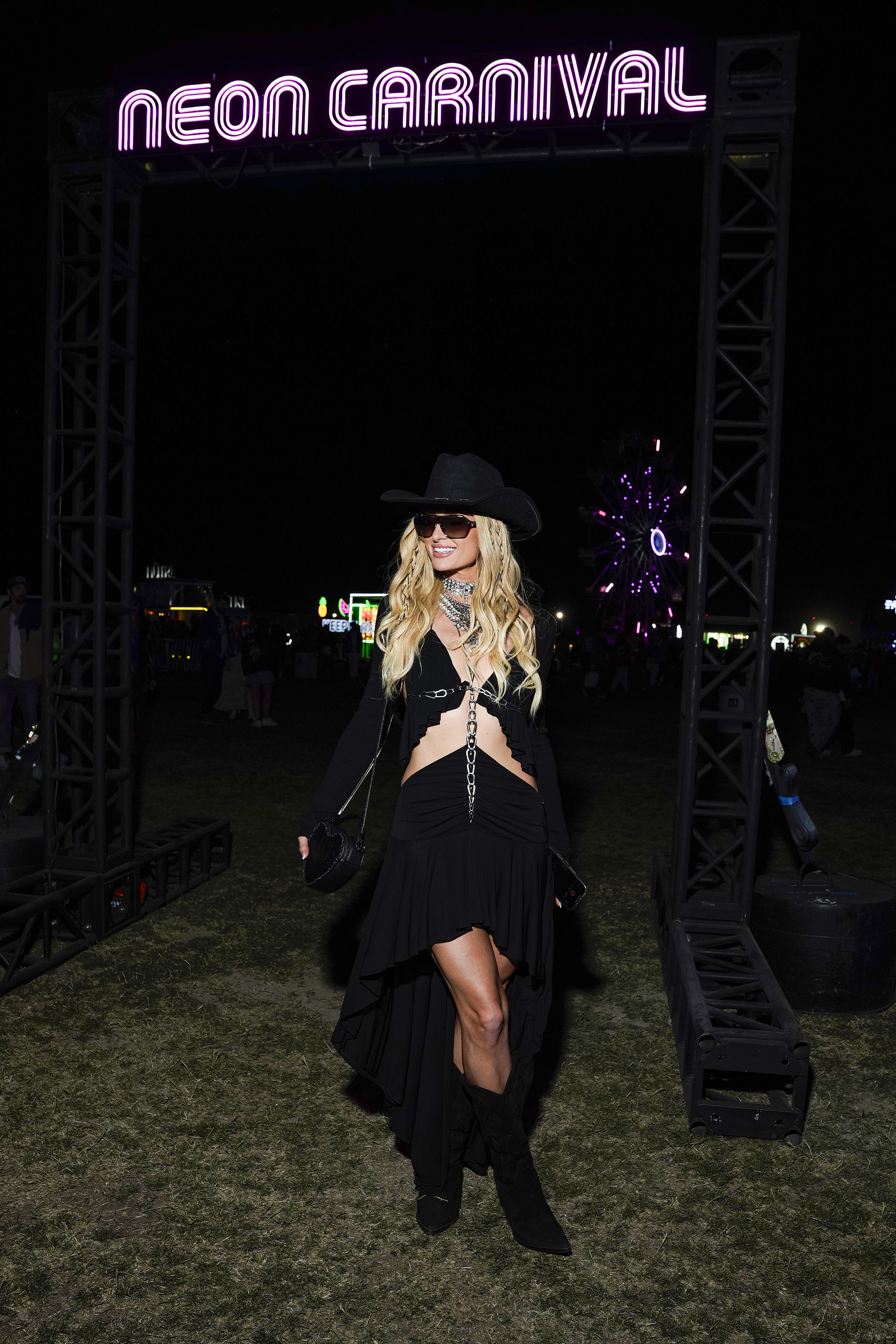 Woman in a black high-slit dress, hat, sunglasses, and boots at Neon Carnival event