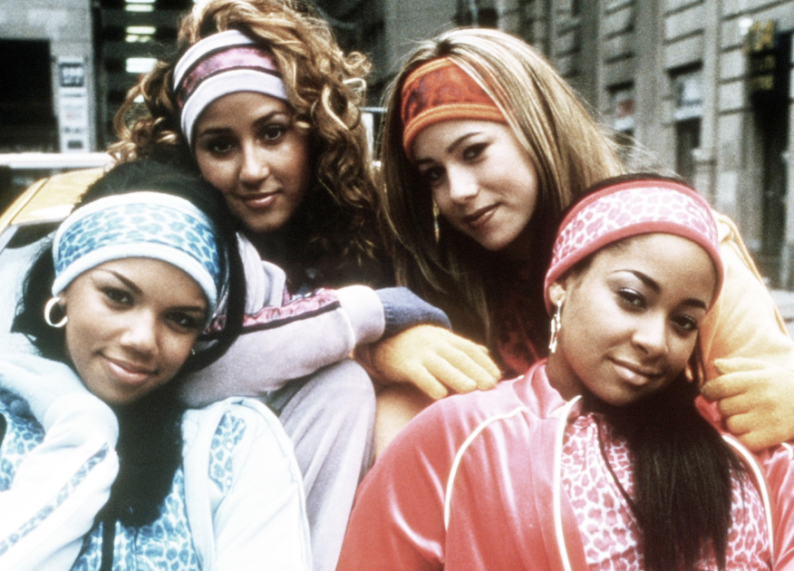 Four women in 90s style clothing and headbands pose together on a city street