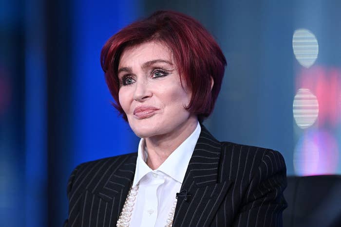 Sharon Osbourne, wearing a pinstripe suit and pearl necklace, is speaking at an event