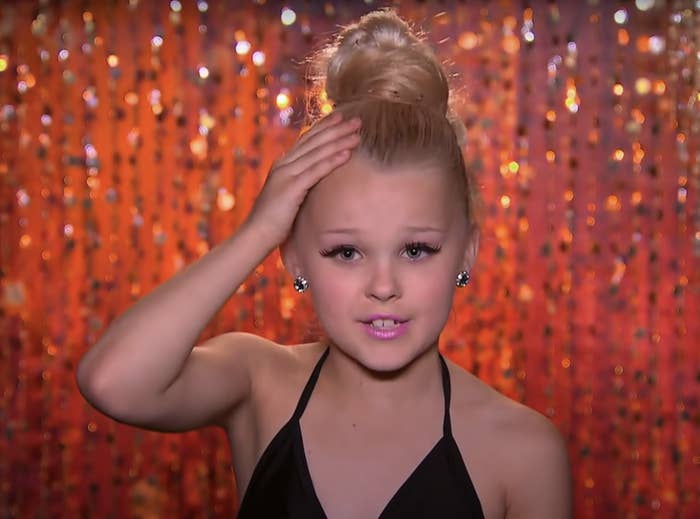 JoJo Siwa wearing a black outfit with hair in a bun, hand on forehead, against a sparkling backdrop