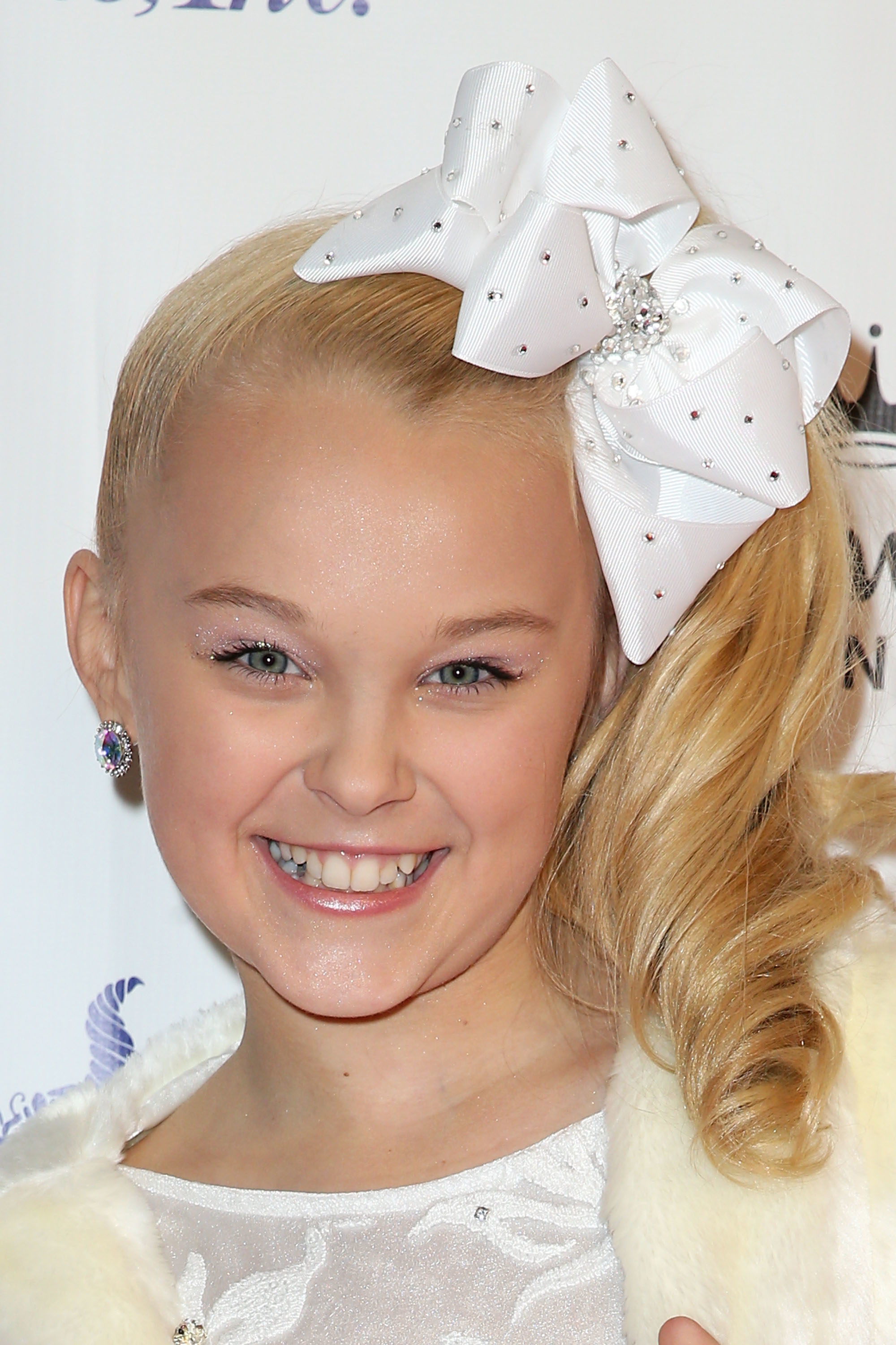 JoJo Siwa smiling, wearing a large bow, earrings, and a faux fur shawl over a dress