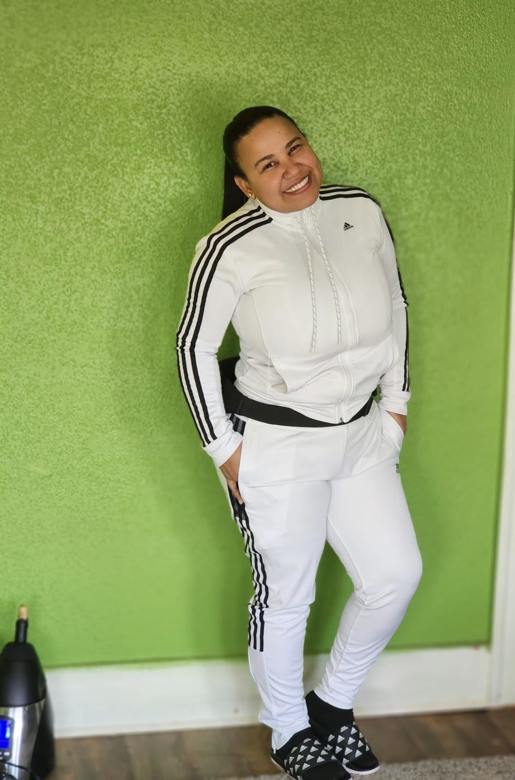 A reviewer in a white Adidas tracksuit with stripes, leaning against a green wall, smiling