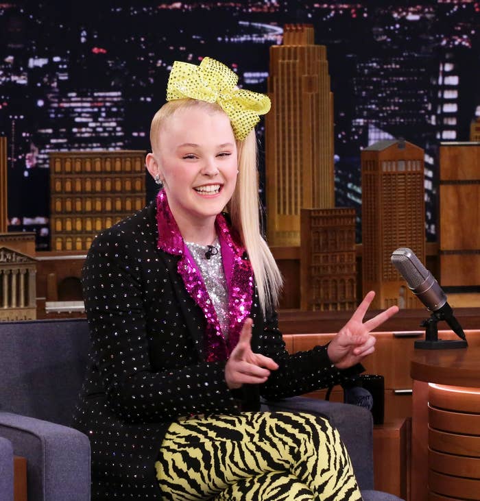 Jojo Siwa seated on a talk show set, wearing a sequined top, animal print pants, large bow, and sneakers