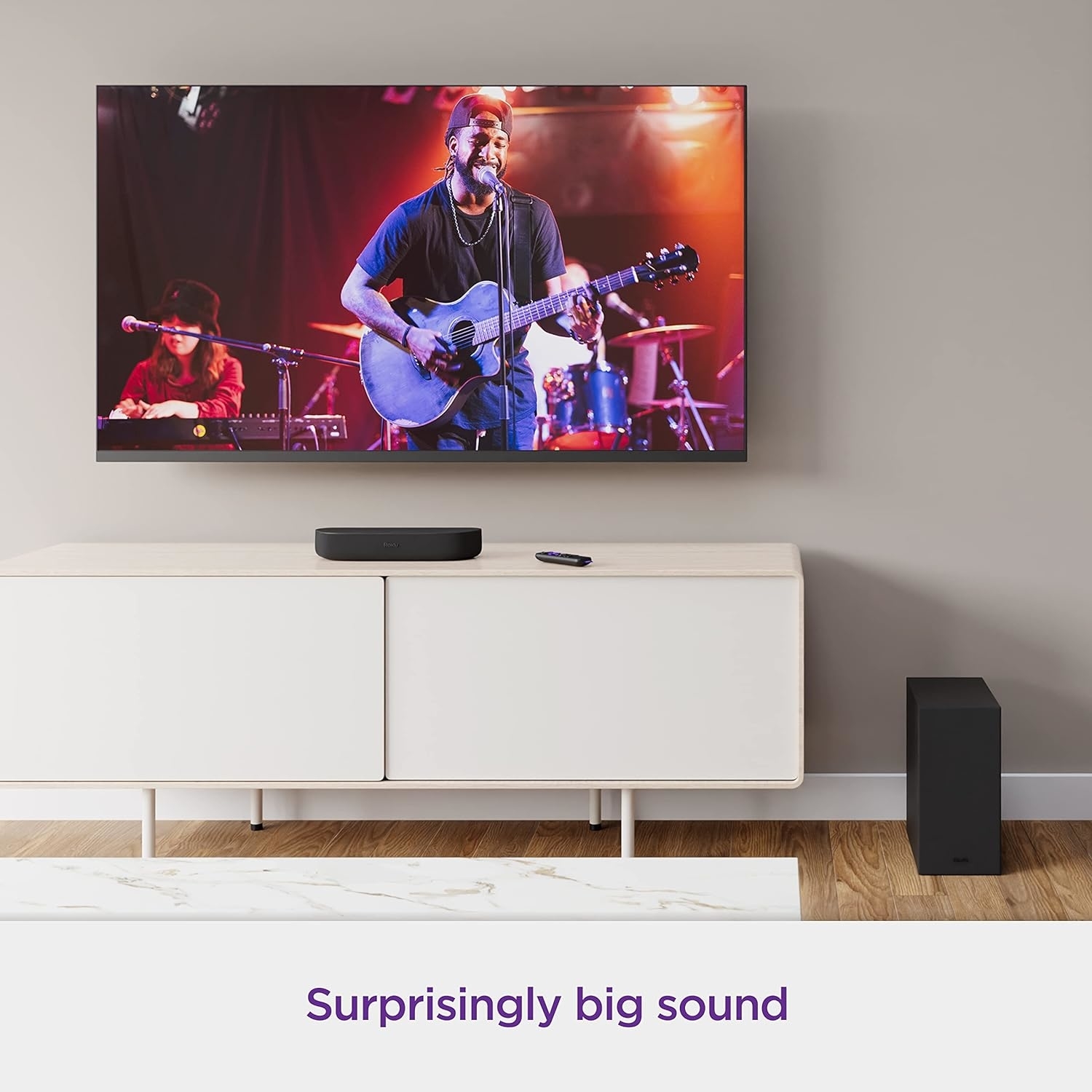 A living room with a TV showing a man playing guitar, the streambar below it, and the subwoofer bass speaker to the side with text &quot;Surprisingly big sound&quot;