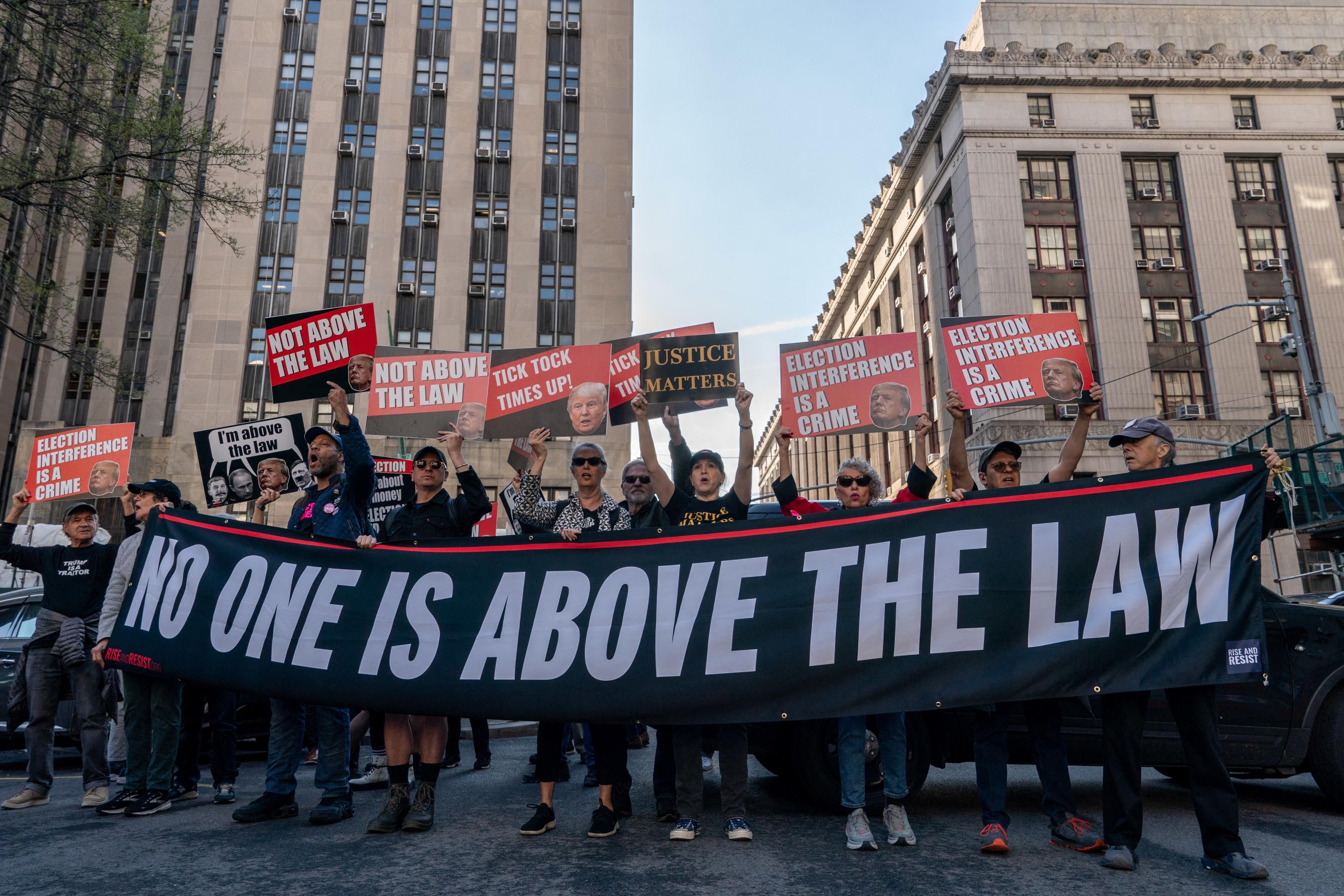 Group of people holding signs and a large banner that reads &quot;NO ONE IS ABOVE THE LAW&quot; during a demonstration