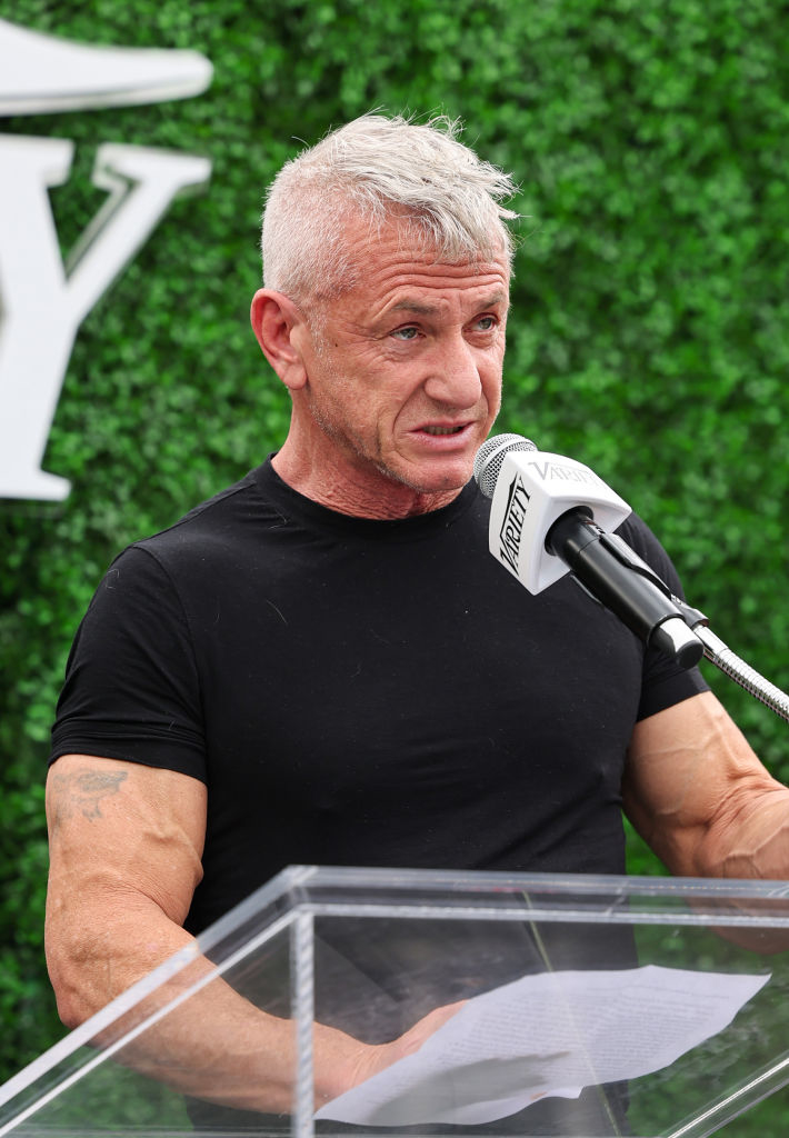 Sean Penn speaking at a microphone, wearing a black short-sleeved top, with a leafy backdrop