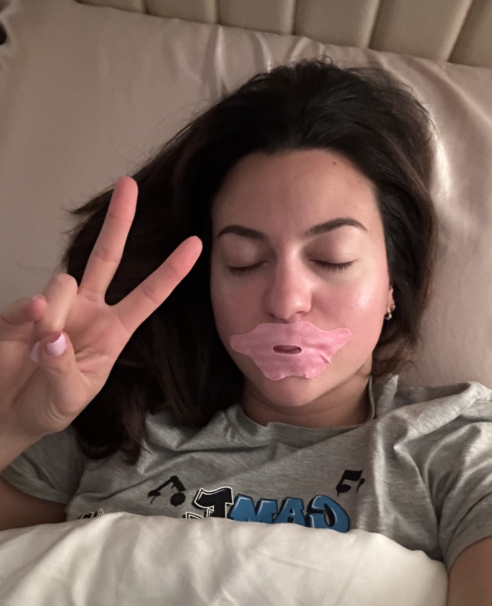 Person in bed with eyes closed, peace sign, wearing lip mask and graphic t-shirt