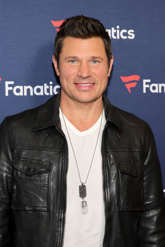 Nick Lachey in a leather jacket and white shirt with a necklace, standing before a promotional backdrop