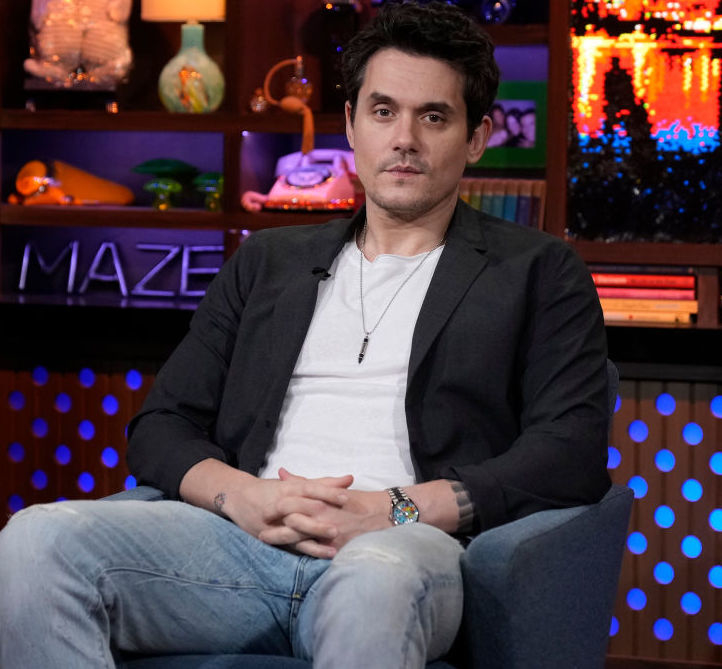 John Mayer sits wearing a blazer over a tee, hands clasped in a TV show setting