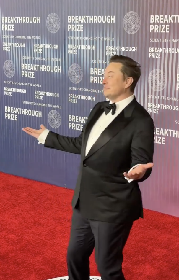 Elon Musk in a tuxedo gesturing, standing in front of a backdrop with &#x27;Breakthrough Prize&#x27; logos