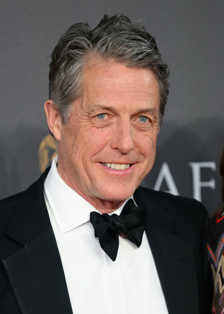 Hugh Grant in a black tuxedo and bow tie smiling at the camera