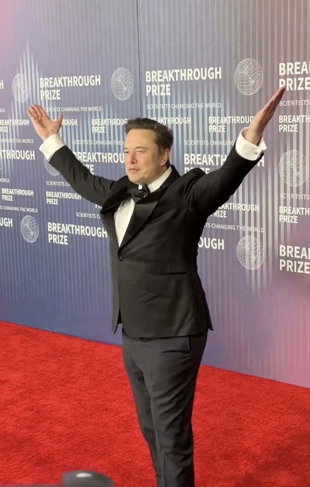 Elon Musk stands with arms outstretched, wearing a black tuxedo at the Breakthrough Prize event