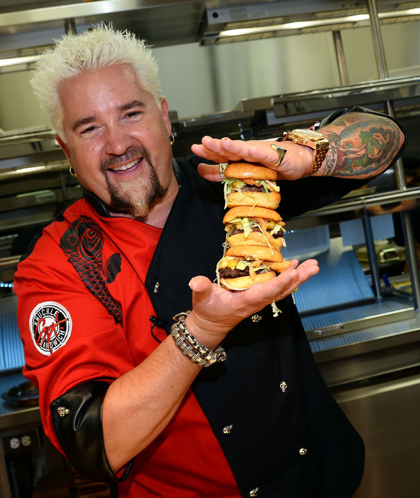 Guy Fieri in a chef&#x27;s outfit, holding a tall stacked burger with multiple layers