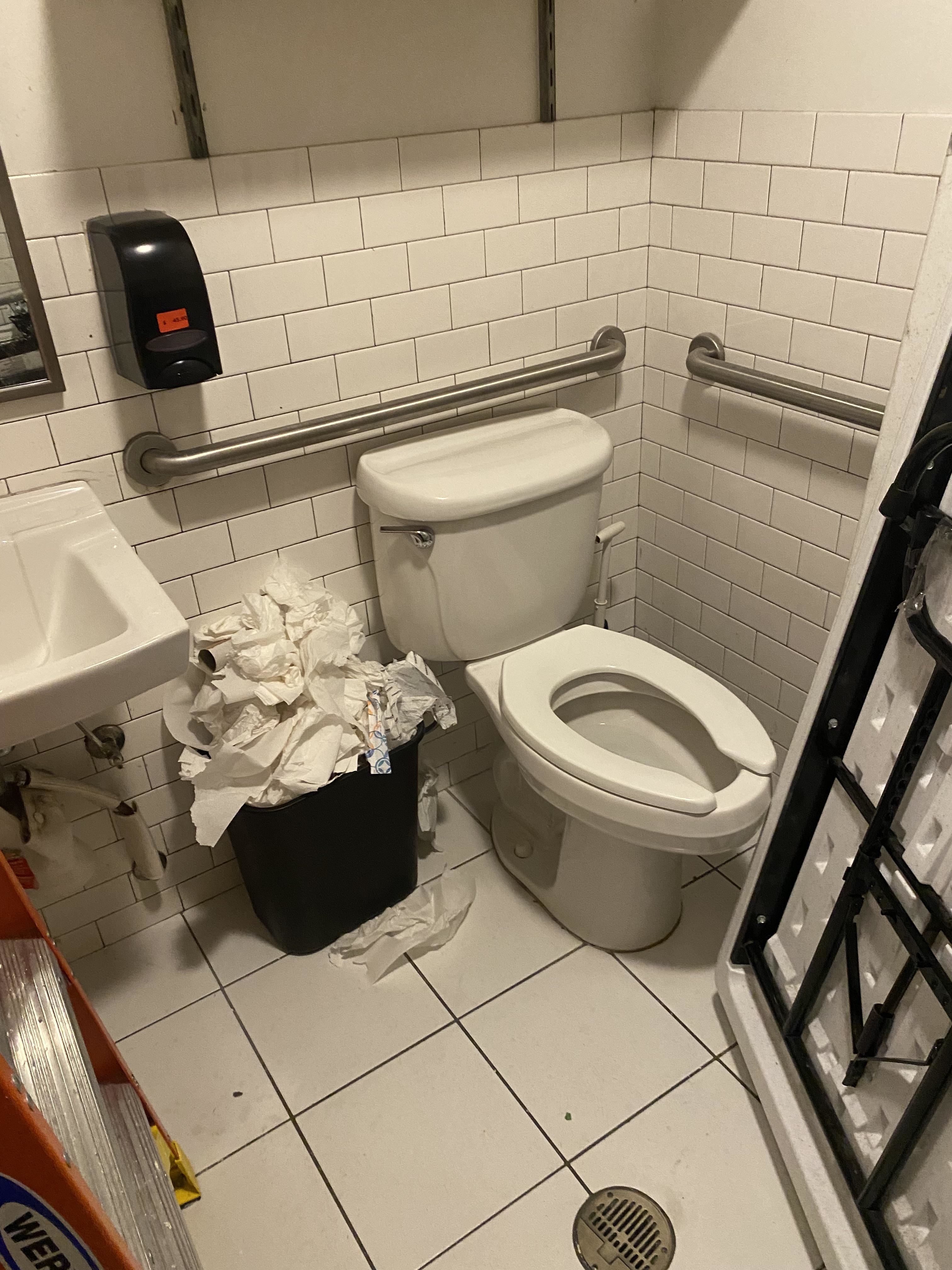 An overflowing bathroom trash can beside a toilet, with paper waste on the floor and a sink nearby