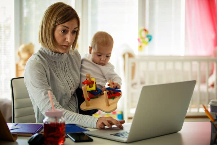 Woman working on a laptop with a baby on her lap holding a toy. They are indoors, suggesting work-life balance for parents