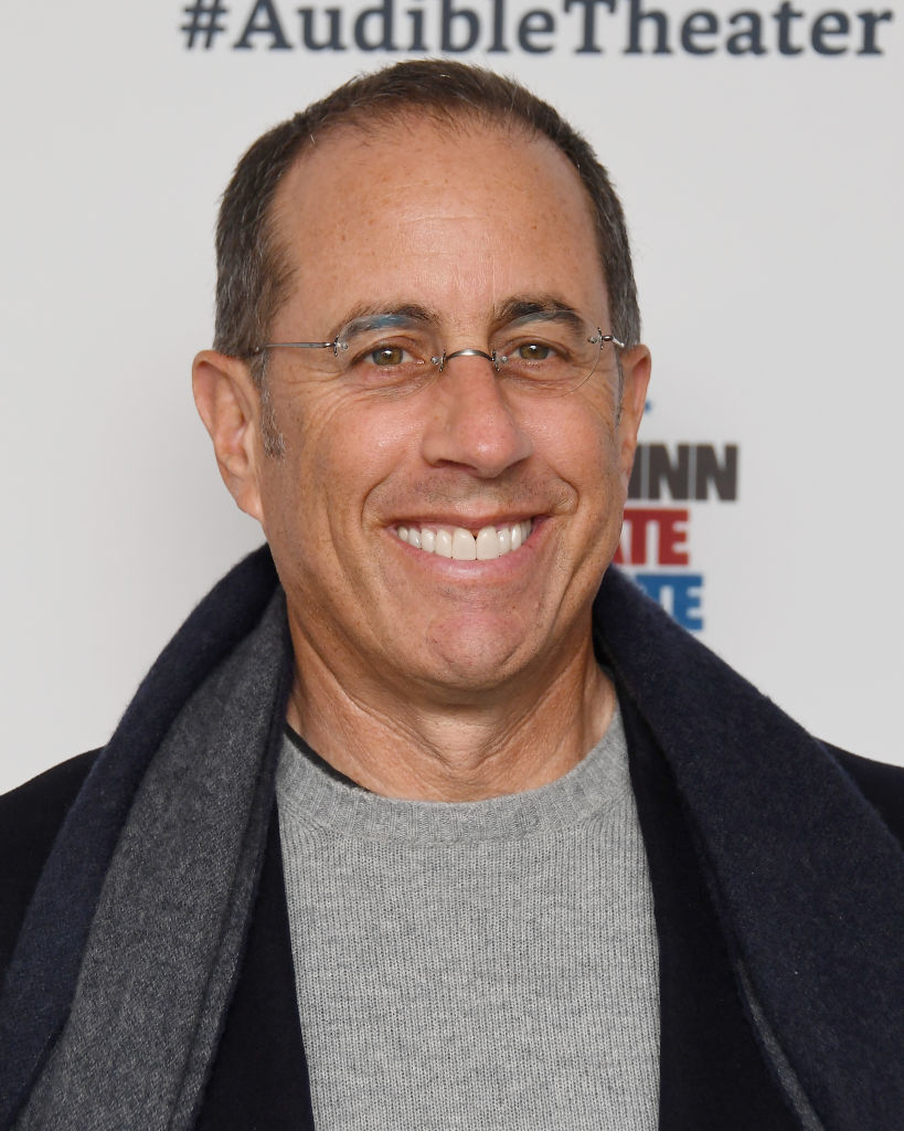 Man smiling at camera, wearing glasses, a grey sweater, and dark coat on shoulders