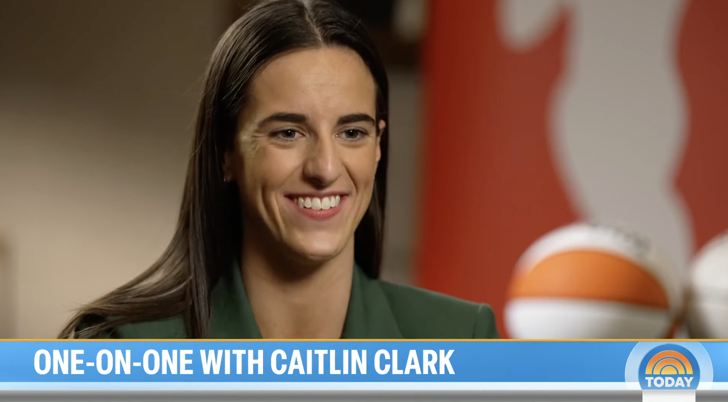 Caitlin Clark in an interview, smiling, wearing a green blazer with basketballs in background, text &quot;ONE-ON-ONE WITH CAITLIN CLARK&quot; below