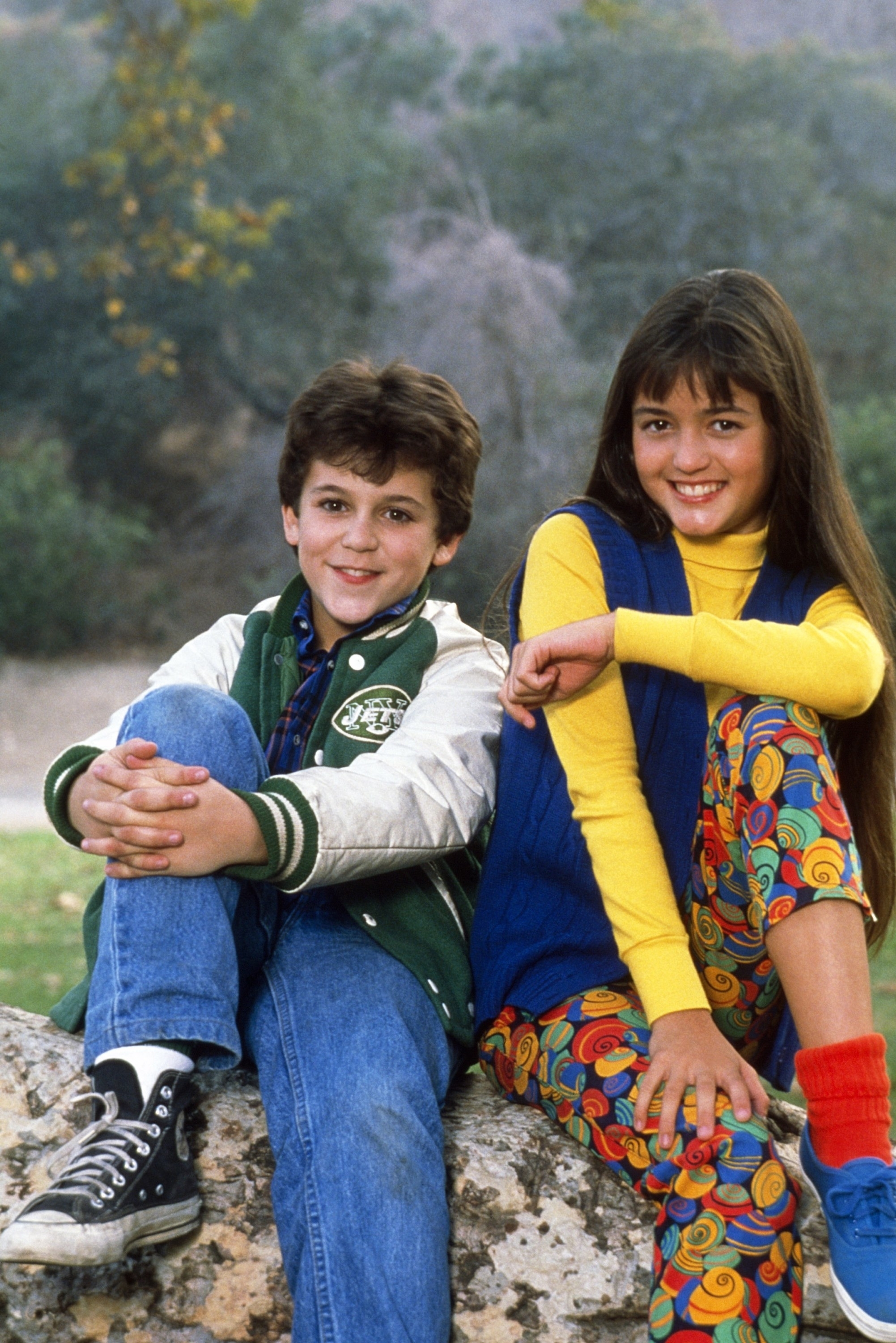 Two young actors from a vintage TV show sitting on a rock, sporting trendy 90s attire