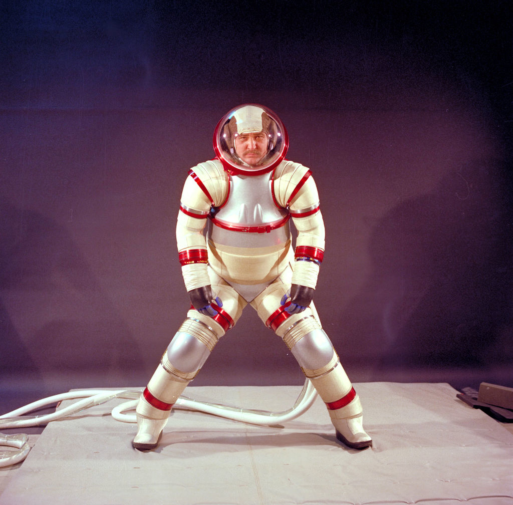 Individual in a vintage spacesuit with helmet and tubes, standing against a plain backdrop