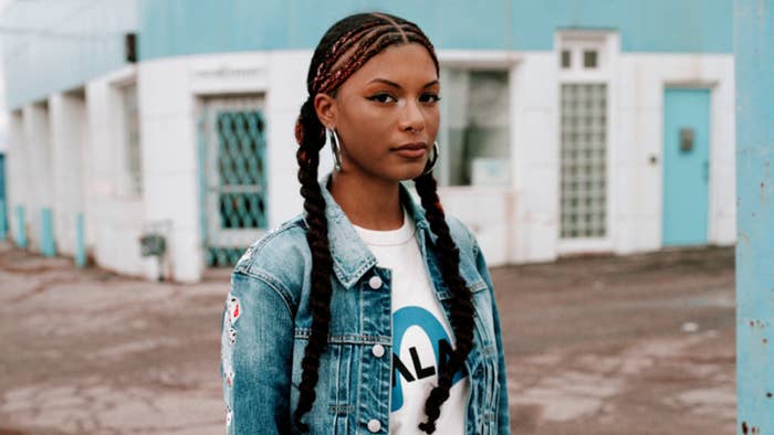 Woman in a graphic tee and denim jacket with braided hair standing before a building