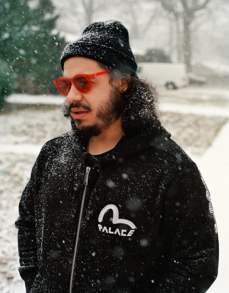 Man in a beanie and sunglasses stands with snow falling around him, wearing a branded hoodie