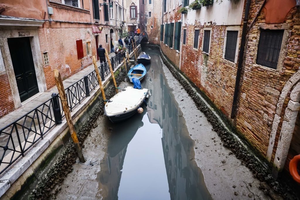 Narrow Venice canal with moored boats and buildings on either side. A person stands by the canal