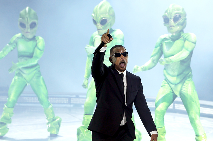 Performer in suit with mic on stage; two dancers in alien costumes behind. Used for music article context