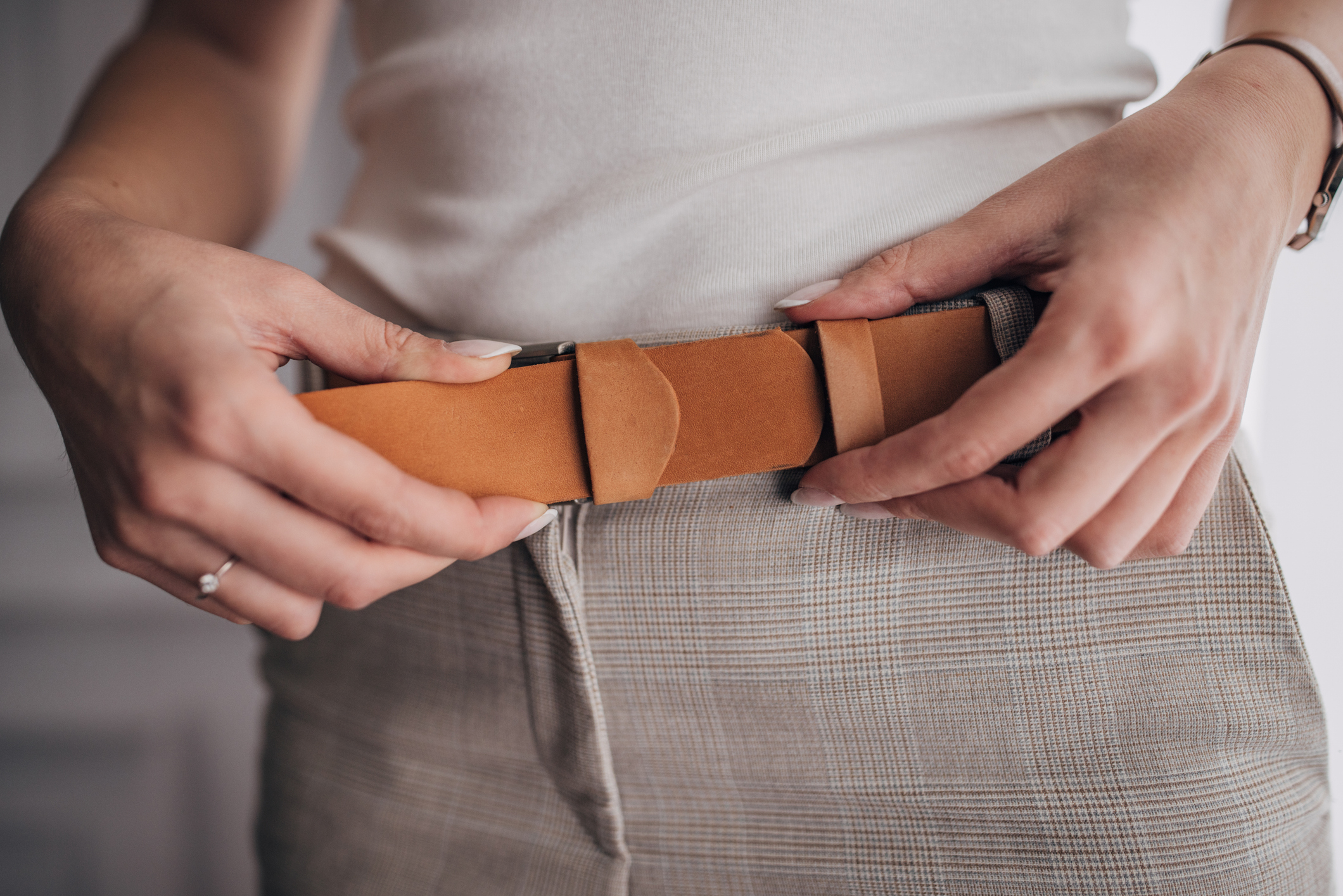 Person fastening a brown leather belt on gray trousers, symbolizing budget tightening or financial planning