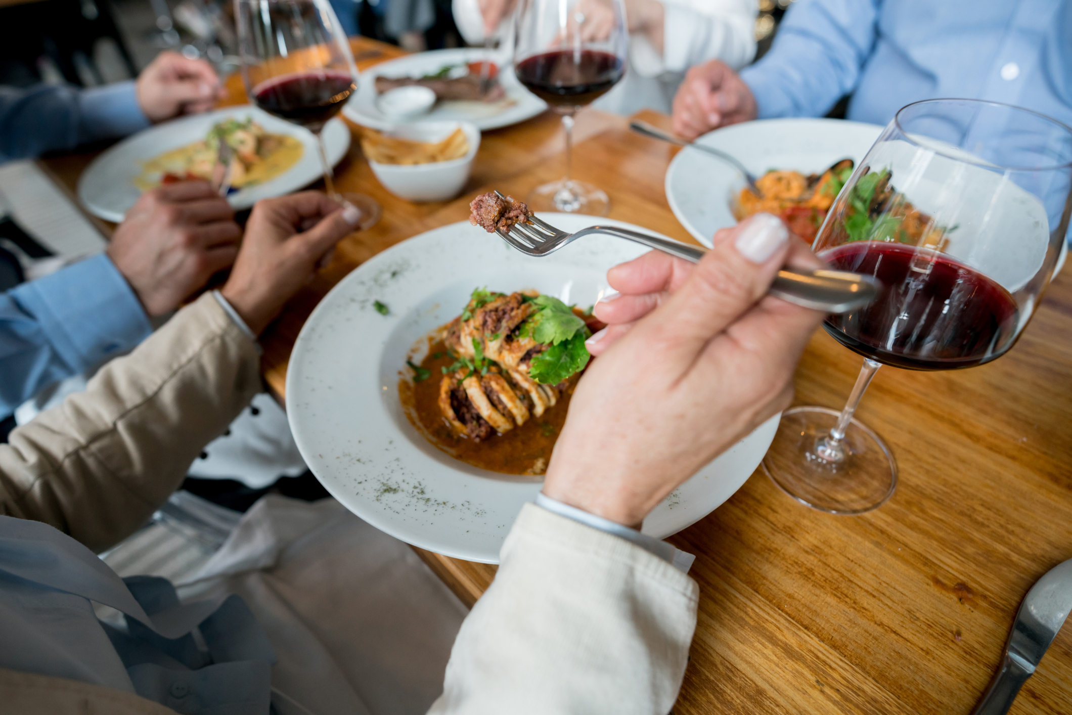 People dining at a table, close-up of hands, plates with food, and glasses of red wine