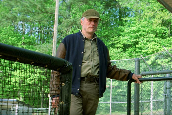 Clint Eastwood in casual attire with a cap leaning on a fence, outdoor setting